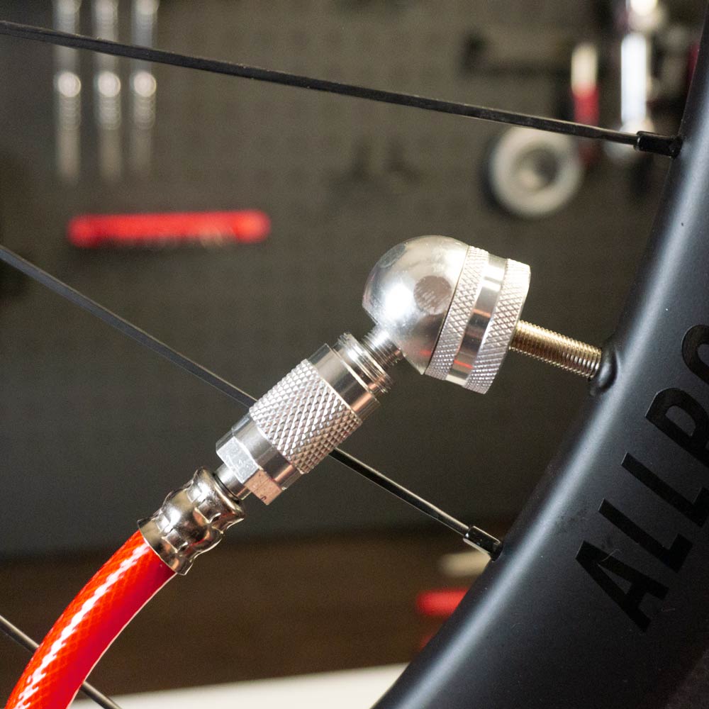 A close-up of the Prestacycle Big Angle Presta Head by Prestacycle, with a red air hose and metal nozzle attached to a bicycles Presta valve on a black wheel for tubeless tire inflation.