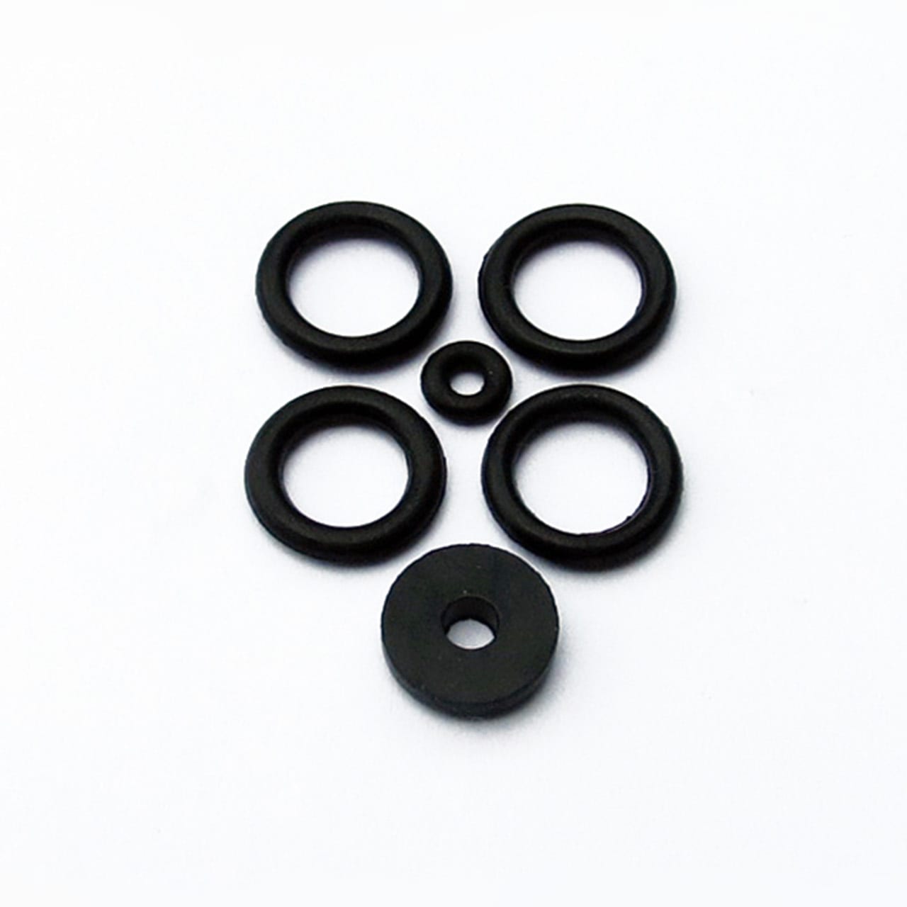 Five black o-rings and one washer from the Prestacycle Prestaflator Rebuild Kit are arranged on a white surface to resemble a simple face, with the washer as the mouth and the smallest o-ring as the nose.