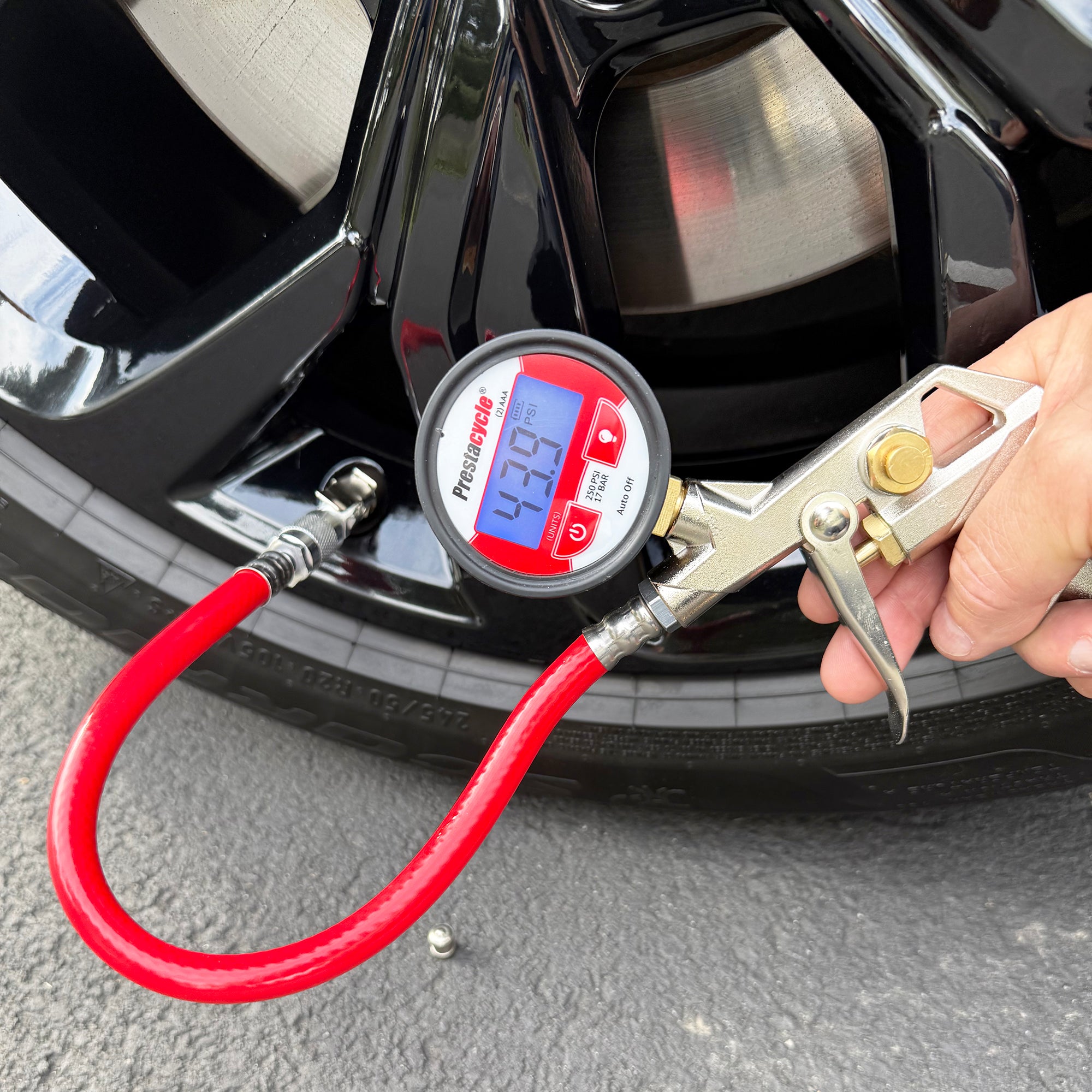 A Prestacycle Prestaflator PRO Digital Bicycle Tire Inflator—compatible with Presta & Schrader valves—is shown attached to a car tire, displaying 42.70 PSI, as a hand holds it against the wheel on a paved surface.
