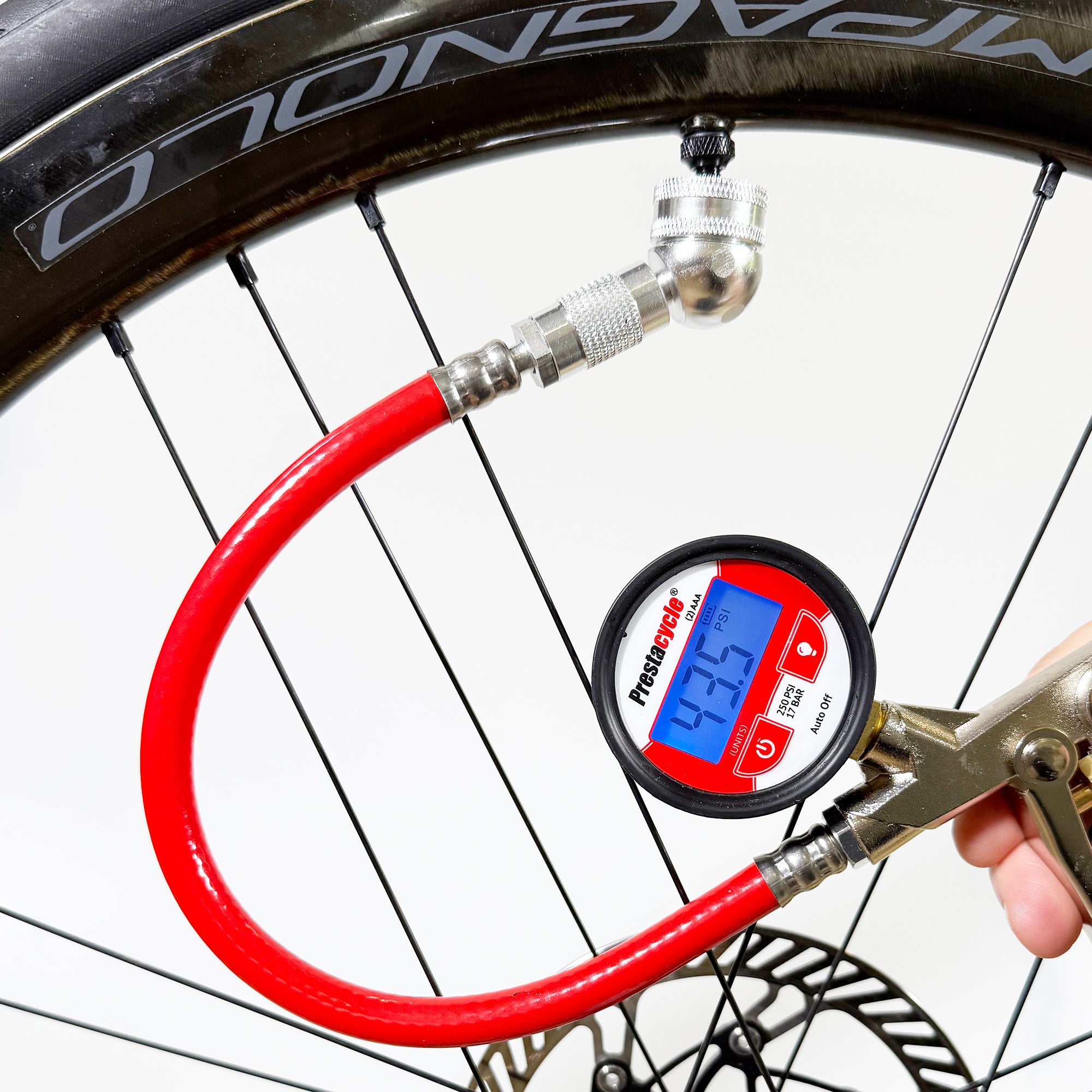 A close-up of a person using the Prestacycle Prestaflator Digital Bicycle Tire Inflator with a red hose to check bicycle tire pressure. The Presta & Schrader air compressor tool displays a reading on its digital screen and is tubeless-safe.
