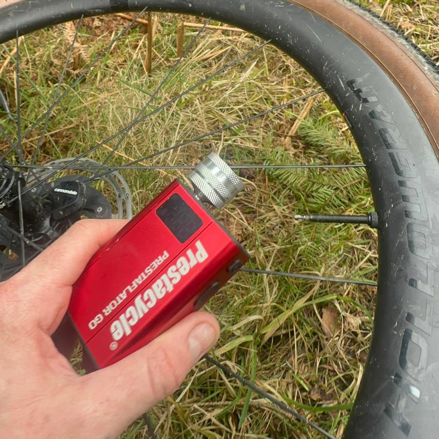A hand holds the Prestacycle Prestaflator GO Portable Digital Rechargeable Preset Bicycle Tire Inflator next to a bike wheel on grass, checking the tire’s air pressure for accurate inflation.