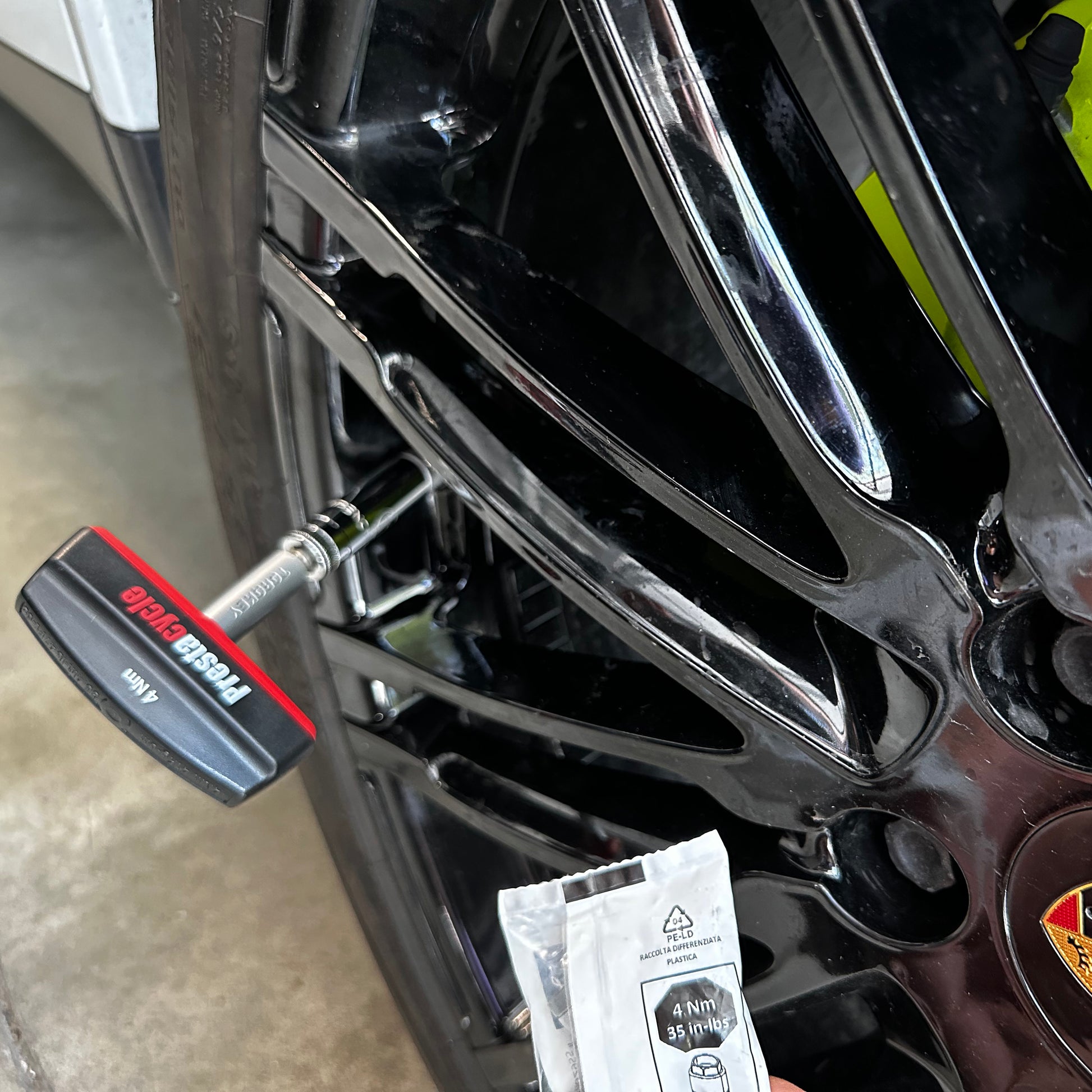 A close-up of a hand holding a packet near a black car wheel, with the Prestacycle Pro TorqKeys 5Nm T-Handle Torque-Limiting Bits Tool by Prestacycle attached to a bolt on the wheel. The Porsche logo appears on the center cap.