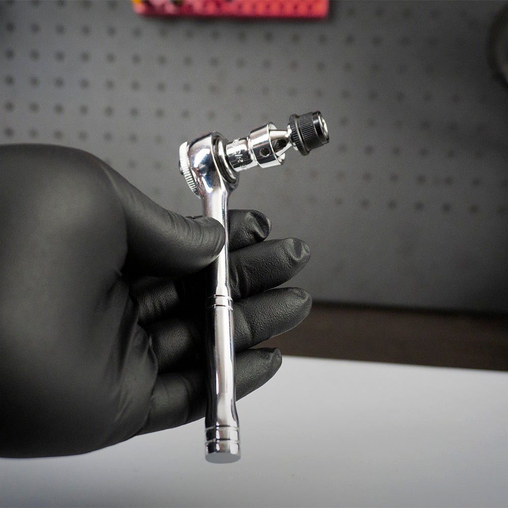A hand in a black glove holds the Prestacycle Bits Swivel QR Torque Wrench Adapter by Prestacycle, a shiny metal tool with socket attachment, against a dark perforated background.