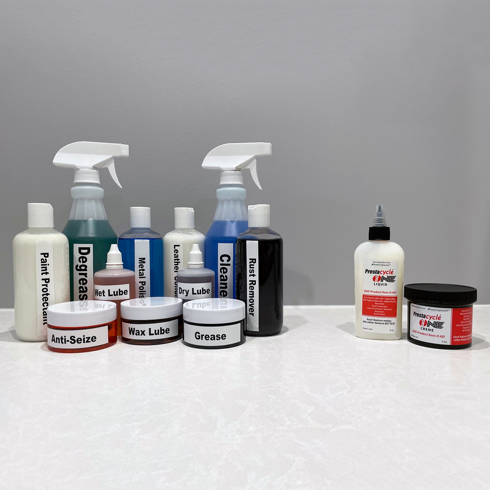 Prestacycle One Liquid and other Prestacycle bike maintenance products, such as lubricant bottles, sprays, degreasers, cleaners, and USDA Certified Bio-Based grease tubs, are displayed on a white surface in front of a gray wall.