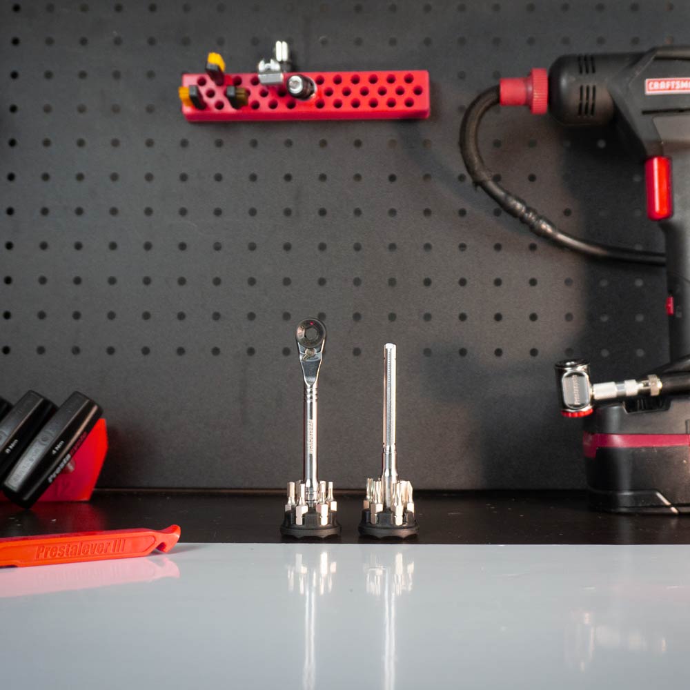 A pegboard wall displays a red power drill, a red tool rack, and various tools—including the Prestacycle Pro T-Handle Kit with 1/4 inch bits by Prestacycle—standing upright on a white workbench in the foreground.