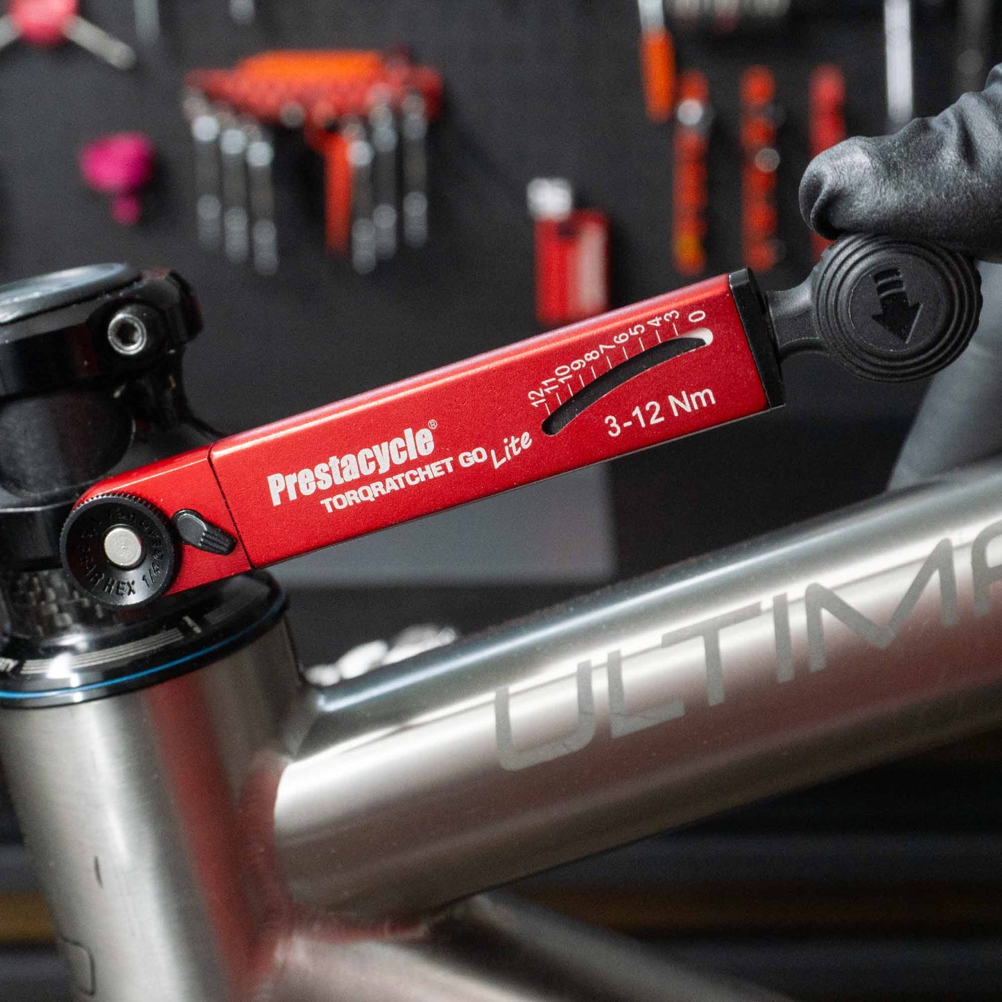 A gloved hand uses a Prestacycle TorqRatchet GO Lite—Portable 3~12Nm Torque Multi-tool Ratchet—on the headset of a silver “ULTIMA” bicycle, with tools hanging on a blurred wall in the background.
