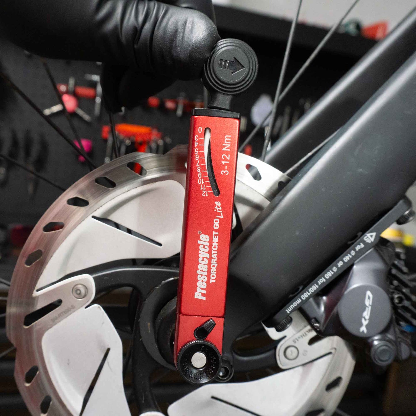 A gloved hand uses a Prestacycle TorqRatchet GO Lite—Portable 3~12Nm Torque Multi-tool Ratchet—to tighten a bolt on a bicycle disc brake rotor. The bike and various tools are visible in the background.