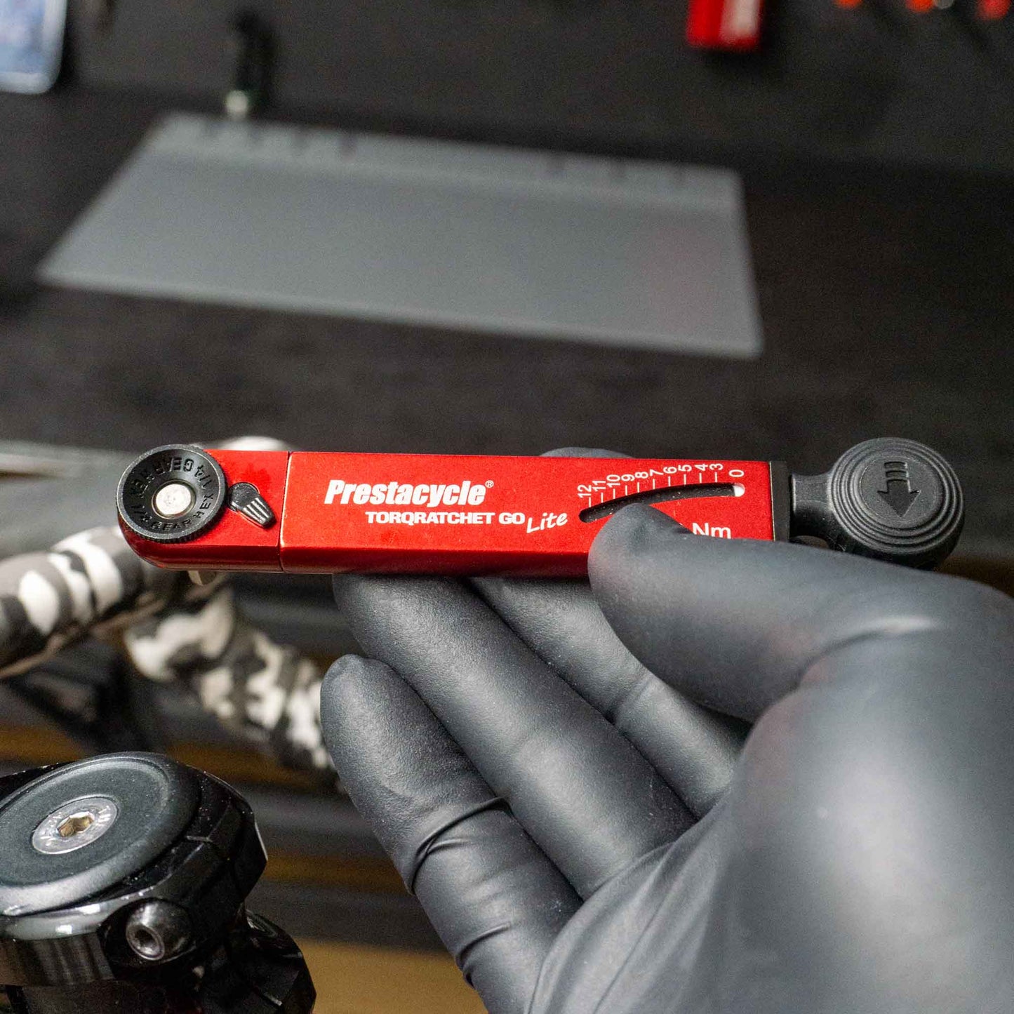 A gloved hand holds a red Prestacycle TorqRatchet GO Lite (3~12Nm) multi-tool ratchet, with a workbench, tools, and bike parts visible in the blurred background.