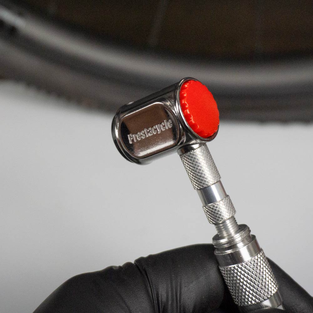 A close-up of a hand in a black glove holding the Prestacycle Pro Presta Head, featuring an auto-lock mechanism and red-capped end, with a blurred bicycle tire in the background.