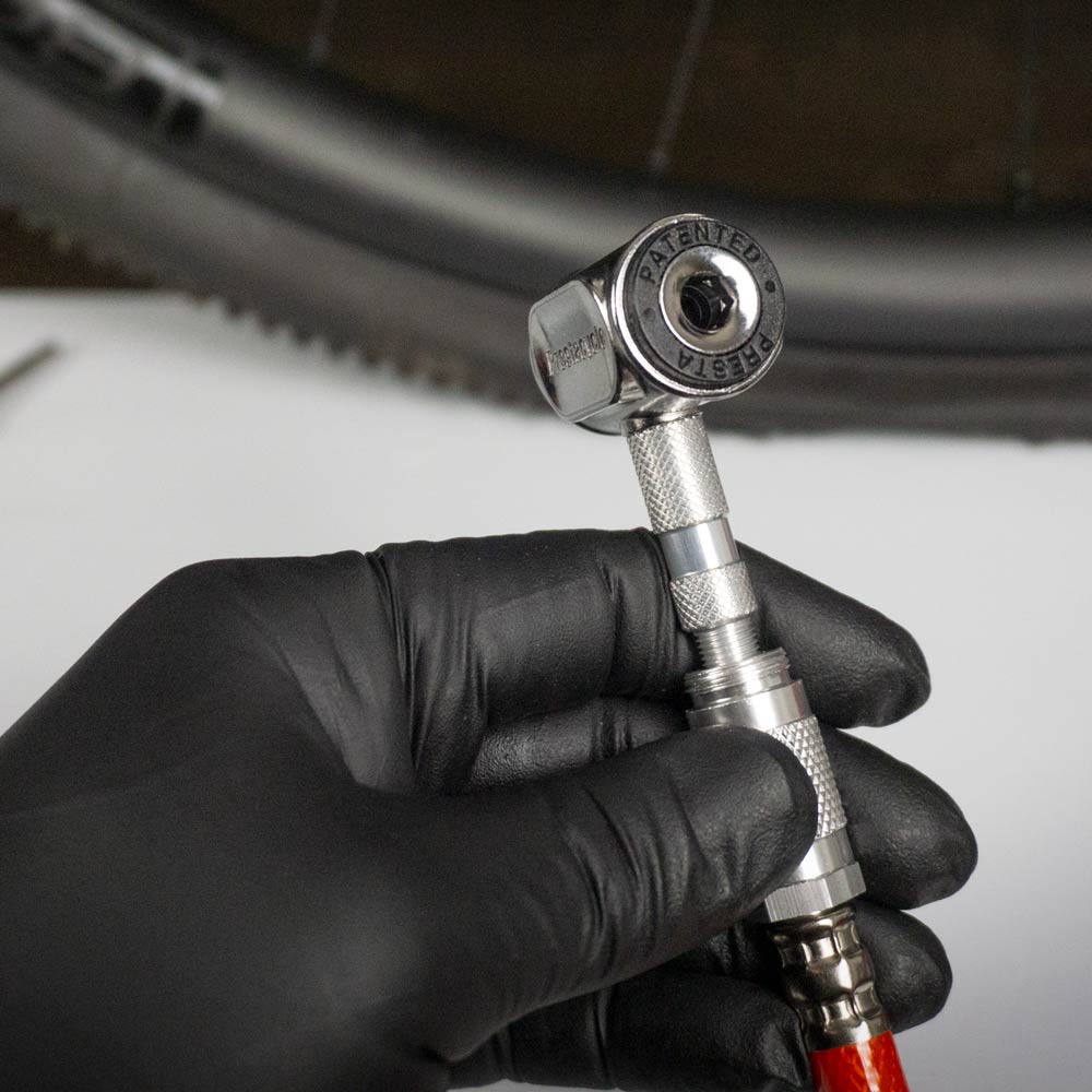 A hand in a black glove holds a Prestacycle Pro Presta Head tire inflator, featuring an instant no-loss disconnect and the Prestacycle brand engraved, positioned near a bike tire in the background.