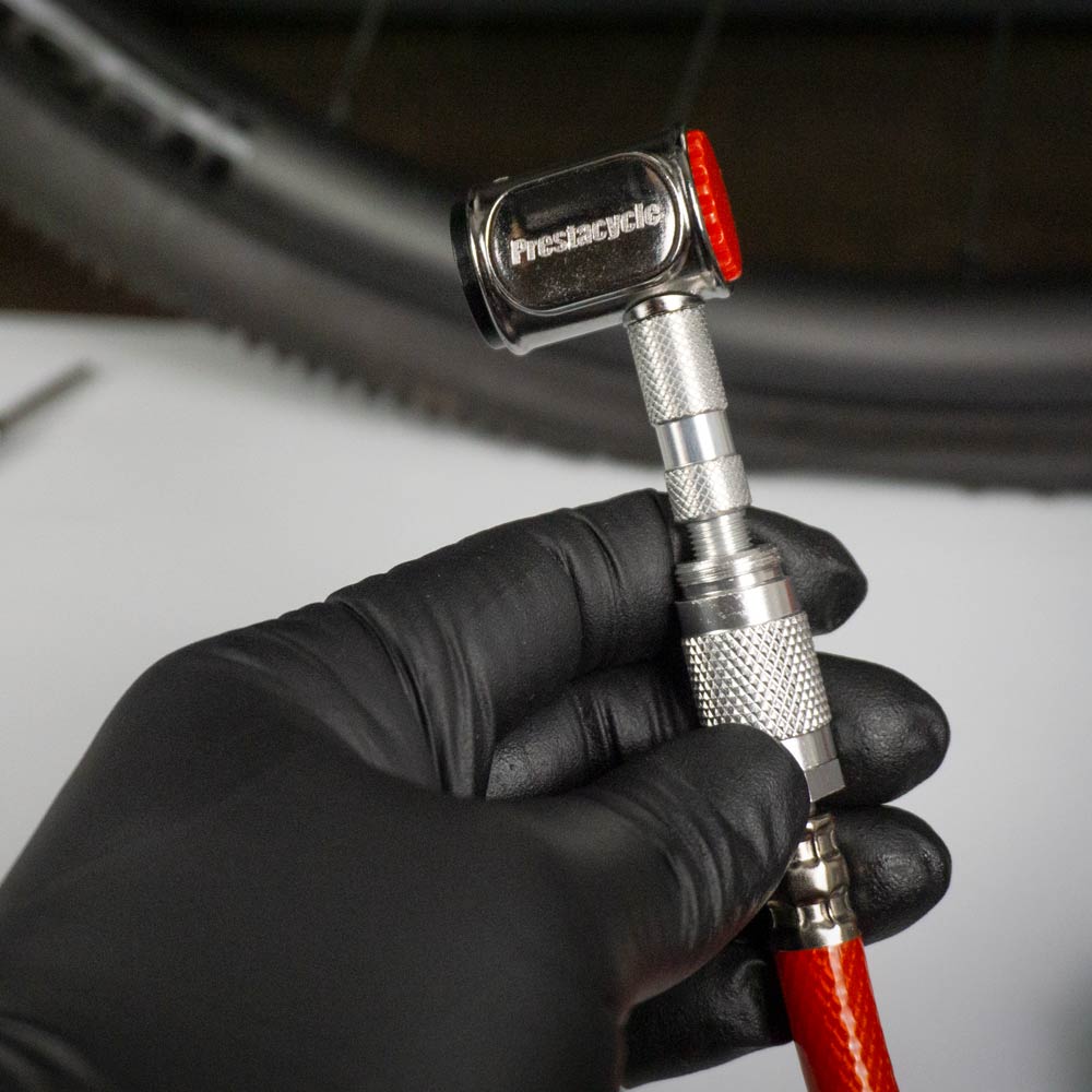 A gloved hand holds a Prestacycle Prestaflator PRO Bicycle Tire Inflator, compatible with Presta & Schrader valves, attached to a red air hose, with a bicycle tire in the background.
