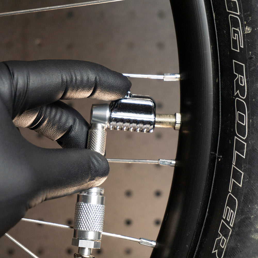 A gloved hand attaches a Prestacycle Pro Schrader Head by Prestacycle with an auto-lock mechanism to a bicycle tire valve; the sidewall shows the words BIG ROLLER.
