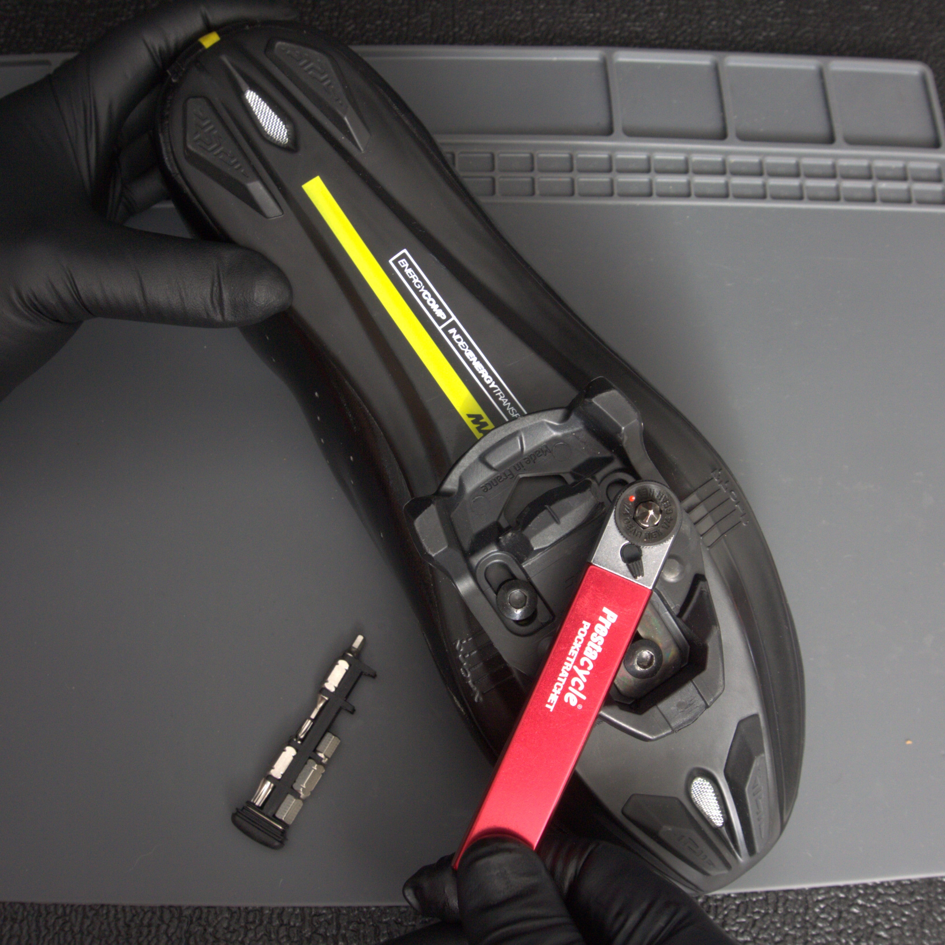 A person wearing black gloves uses the Prestacycle PocketRatchet multi-tool by Prestacycle to adjust the cleat on a black cycling shoe. Its set of bits, stored in the handle, is placed nearby on a gray surface.