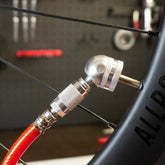A close-up of a Prestacycle Prestaflator Digital Bicycle Tire Inflator - Presta & Schrader Air Compressor Tool attached to a bike valve, with tools blurred in the background.