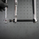 A gloved hand uses a Prestacycle 1/4” Square to Hex Bits Quick Release Adapter to tighten a bolt on a bicycle axle, with part of the bike frame and the adapter visible against a gray, perforated background.
