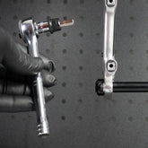 A gloved hand uses the Prestacycle 1/4” Square to Hex Bits Quick Release Adapter near a silver bike crank arm, ready to adjust a bolt. The dark, perforated background is ideal for 1/4″ socket-type torque wrenches.