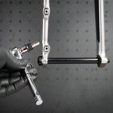 A gloved hand holds the Prestacycle 1/4” Square to Hex Bits Quick Release Adapter near a bicycle frame dropout, preparing to install or tighten a thru-axle. A black pegboard is visible in the background.
