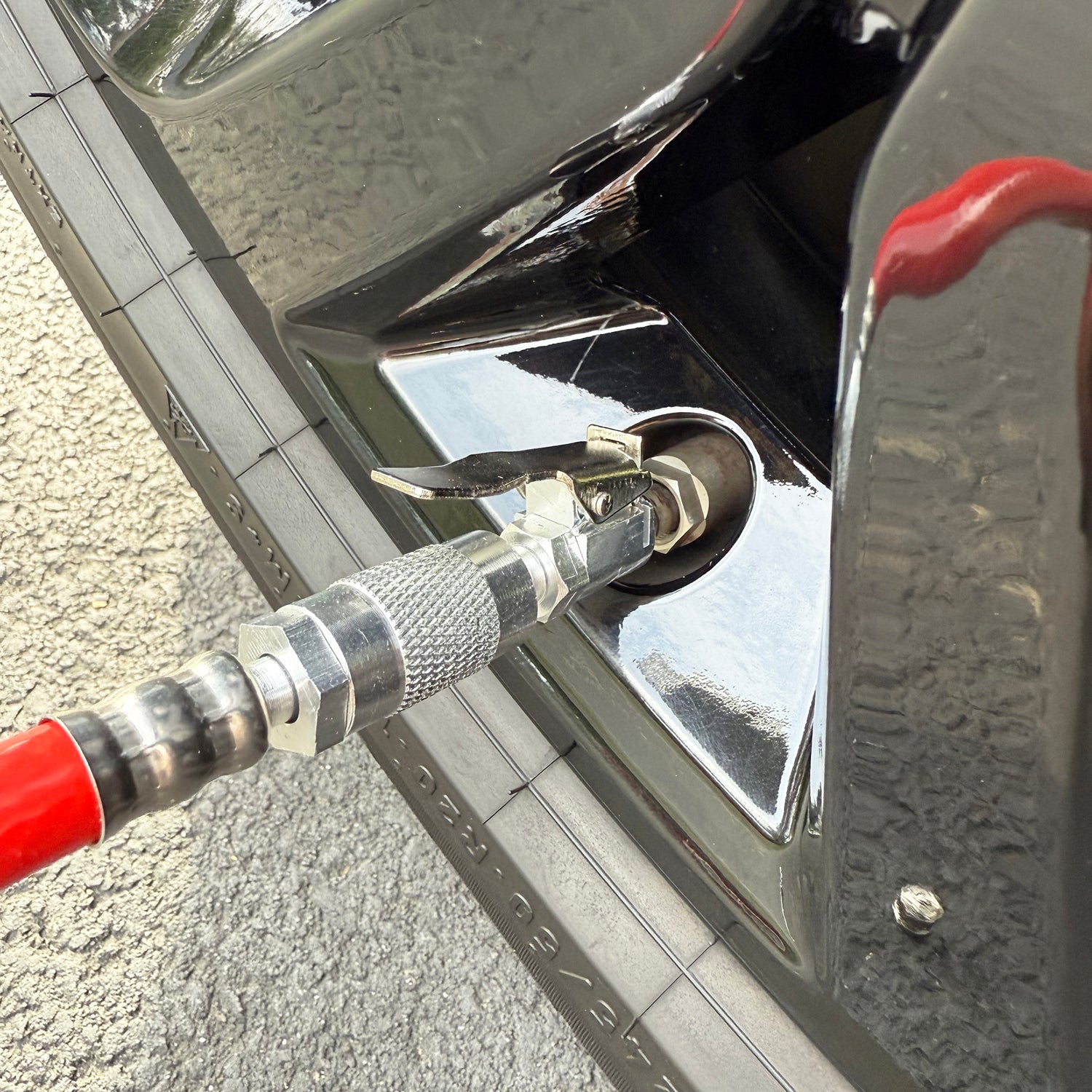 A close-up of a car tire being inflated with the Prestacycle Prestaflator PRO Digital Bicycle Tire Inflator (Presta &amp; Schrader), attached to the valve stem. The car’s shiny black finish and part of the textured tire are visible.