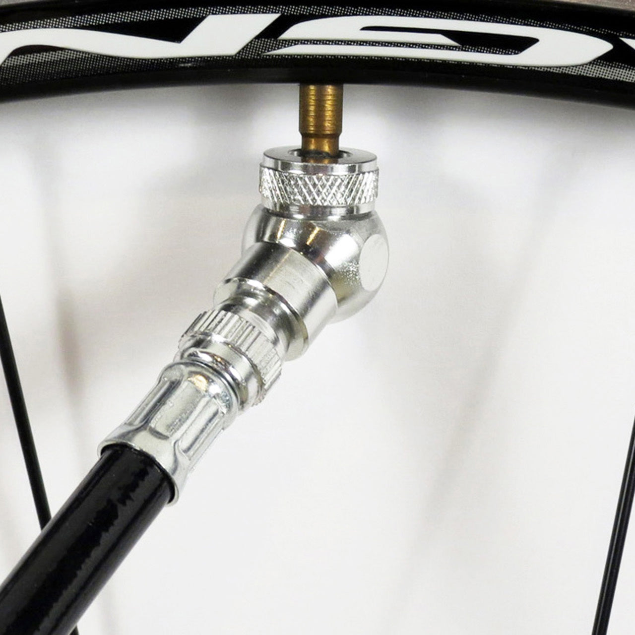 A close-up of a bicycle tubeless tire valve being inflated with the Prestacycle Angle Mini Presta Head pump nozzle attached to the valve stem. Black wheel spokes are visible in the background.