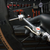 A gloved hand uses the Prestacycle Prestaflator Mini Presta Air Compressor Tire Inflator to inflate a brown sidewall bike tire, with assorted bicycle tools visible on a blurred pegboard in the background.