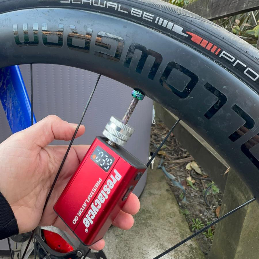 A hand holds a Prestacycle Prestaflator GO - Portable Digital Rechargeable Preset Bicycle Tire Inflator attached to a bicycle valve for precise inflation. The Schwalbe Pro One logo is visible on the black tire as the display shows the reading.