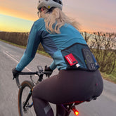 A cyclist with long blond hair, in a teal jacket and helmet, rides at sunset on a rural road. The jacket’s back pockets hold a phone, an energy bar, a gel, and the Prestacycle Prestaflator GO Portable Digital Rechargeable Bicycle Tire Inflator.