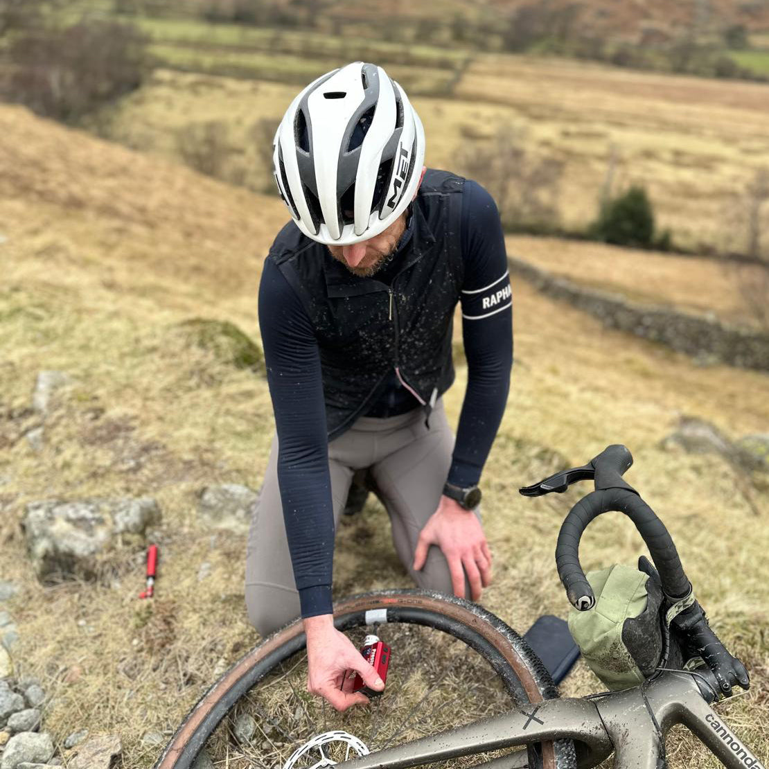 A cyclist in Rapha gear kneels on grass to fix a tire using the Prestacycle Prestaflator GO, a portable digital rechargeable preset bicycle tire inflator, with hills and fields creating a tranquil rural backdrop.