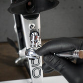A person in a black glove uses an airbrush near a Colnago bike frame, while the Prestacycle Bits Swivel QR Torque Wrench Adapter by Prestacycle sits nearby, ready for precise adjustments after custom paintwork.
