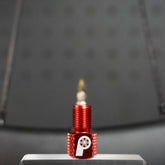 The Prestacycle Prestadapter TUBELESS, a red lightweight alloy Presta adapter and valve core remover with a white wheel logo, stands upright on a silver surface against a blurred, dark background.