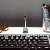 A plain gray background displays a desk with a black keyboard, sunglasses, a glass with pens and a trophy, and the Prestacycle Prestaratchet Micro Kit (6 S2 bits, round holder stand) arranged upright at the center.