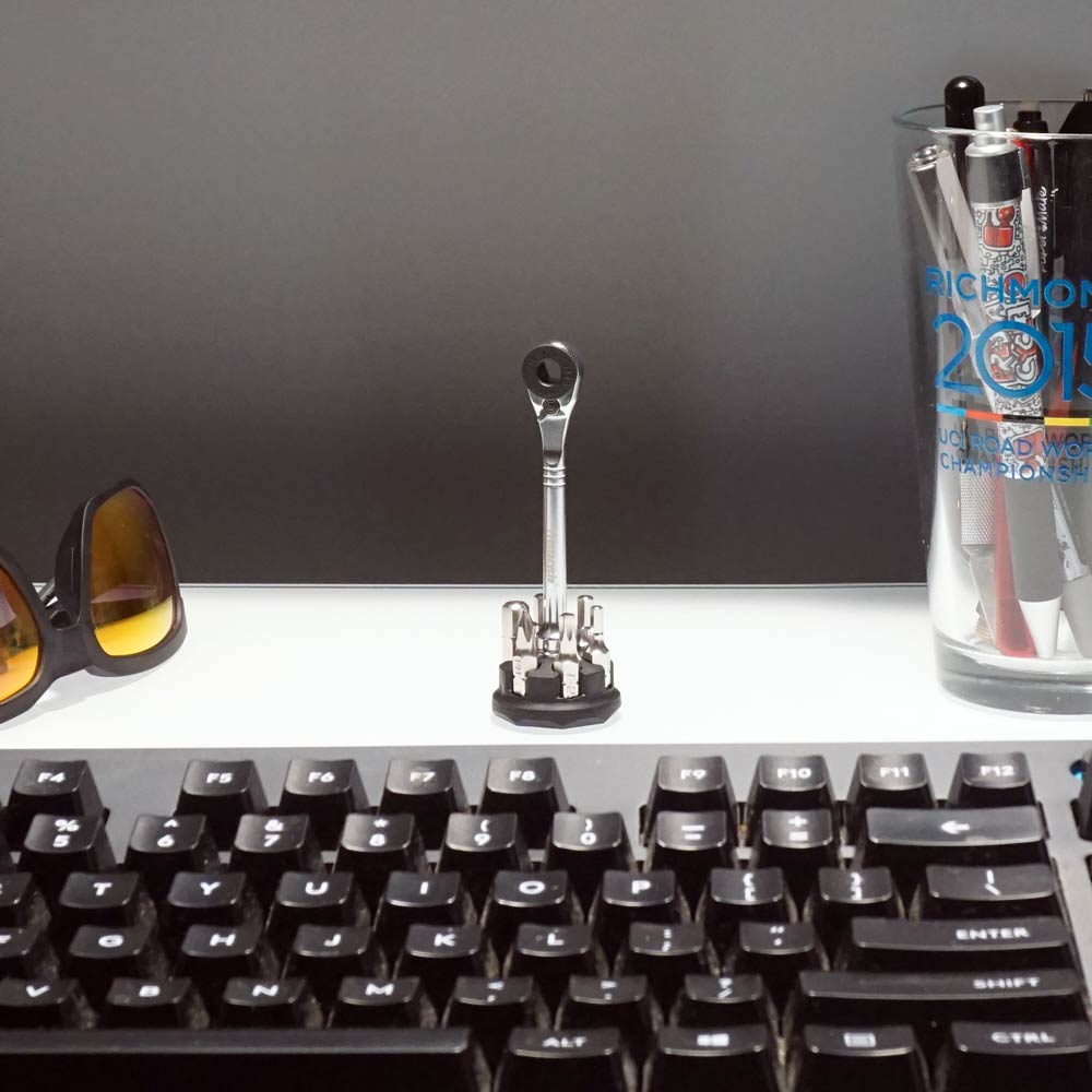 A plain gray background displays a desk with a black keyboard, sunglasses, a glass with pens and a trophy, and the Prestacycle Prestaratchet Micro Kit (6 S2 bits, round holder stand) arranged upright at the center.