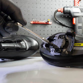 A gloved hand uses a screwdriver to attach a cleat to a cycling shoe, with the Prestacycle Prestaratchet Micro Kit (includes 6 S2 bits and round holder stand) on the workbench in the background.