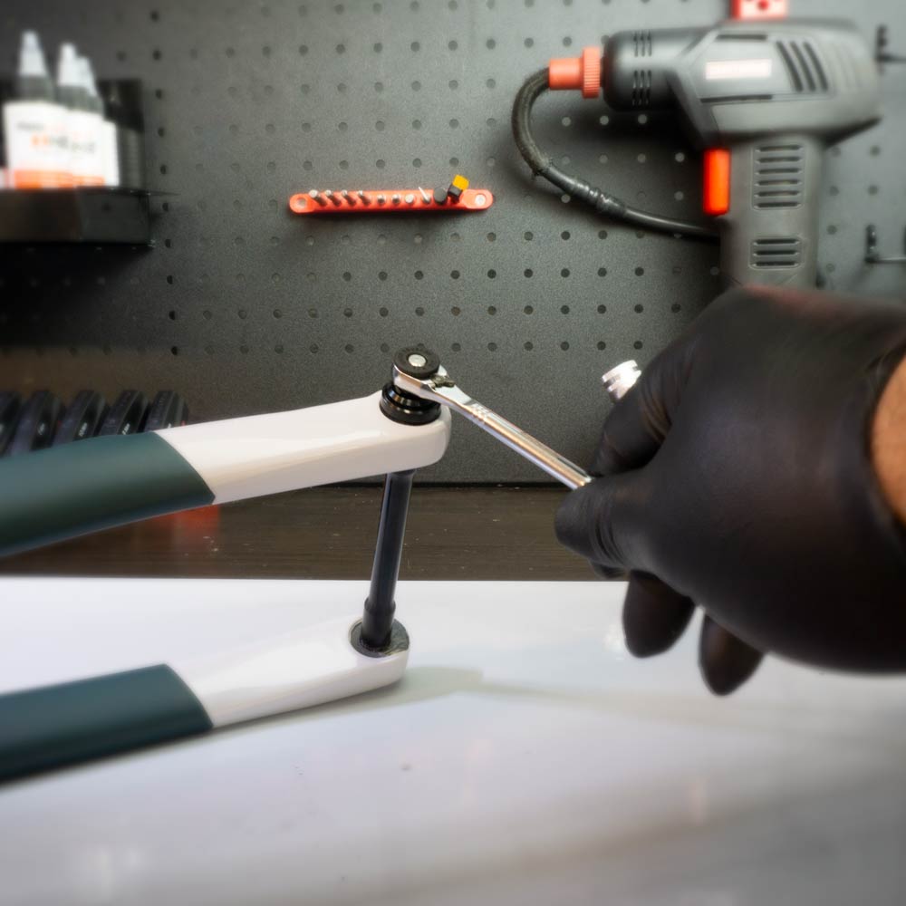 Wearing a black glove, a person uses the Prestacycle Prestaratchet Micro – Kit with (6) S2 Bits and Round Holder Stand to tighten a bolt on a white and green bike fork, with tools and a drill on a pegboard in the background.