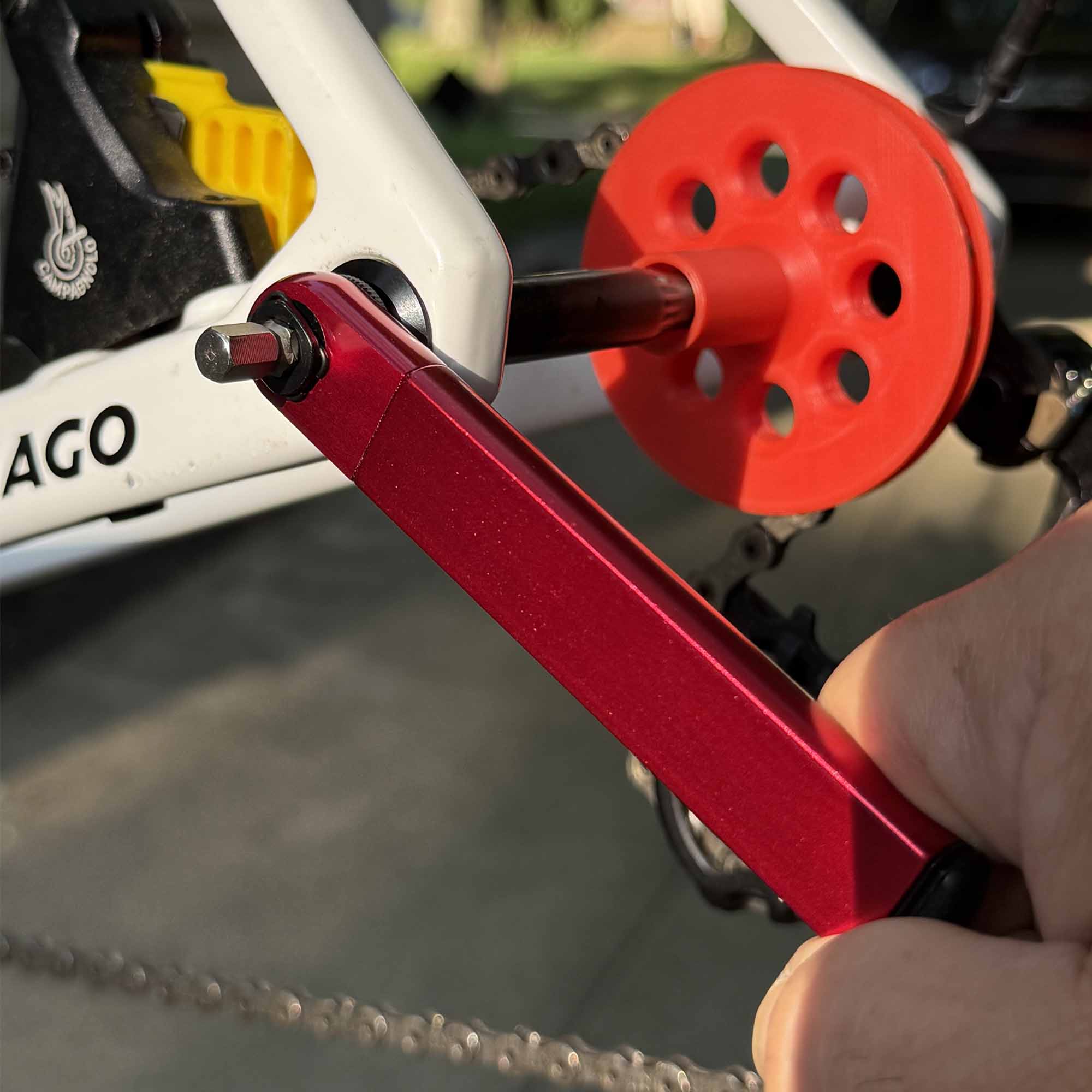 A close-up shows a hand using a red hex wrench to adjust a bike crank, with the Prestacycle PocketRatchet Lite—pocket multi-tool with bits stored in the handle—plus a red plastic disc tool near part of a white bike frame in the background.