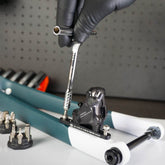 A gloved hand uses the Prestacycle Pro T-Handle Kit to adjust a bicycle disc brake caliper on a bike fork, with other Prestacycle tools and bits from the kit visible nearby on the workbench.