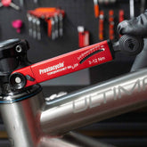 A hand uses a red Prestacycle TorqRatchet GO Lite - Portable 3~12Nm Torque Multi-tool Ratchet to adjust a bolt on a silver ULTIMA bicycle frame, with bike tools hanging on a wall in the blurred background.