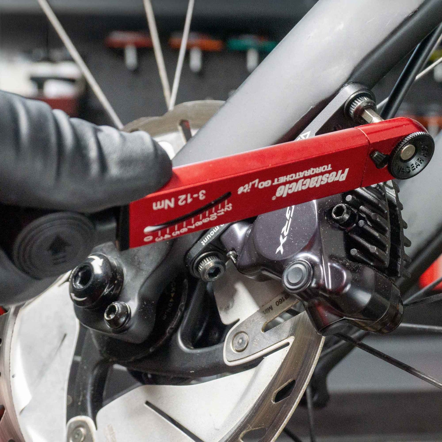 A gloved hand uses the Prestacycle TorqRatchet GO Lite (3~12Nm) to tighten a bolt on a bicycle disc brake caliper, with the bike wheel and brake rotor clearly visible.