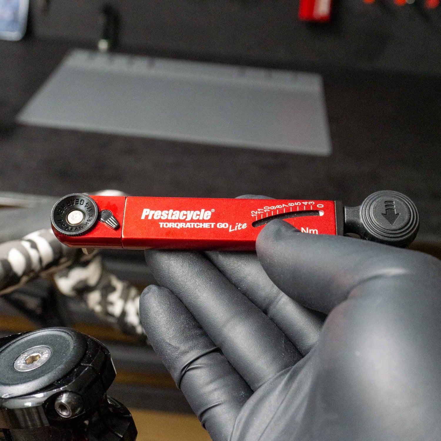 A gloved hand holds a red Prestacycle TorqRatchet GO Lite (3~12Nm) multi-tool ratchet, with a workbench, tools, and bike parts visible in the blurred background.