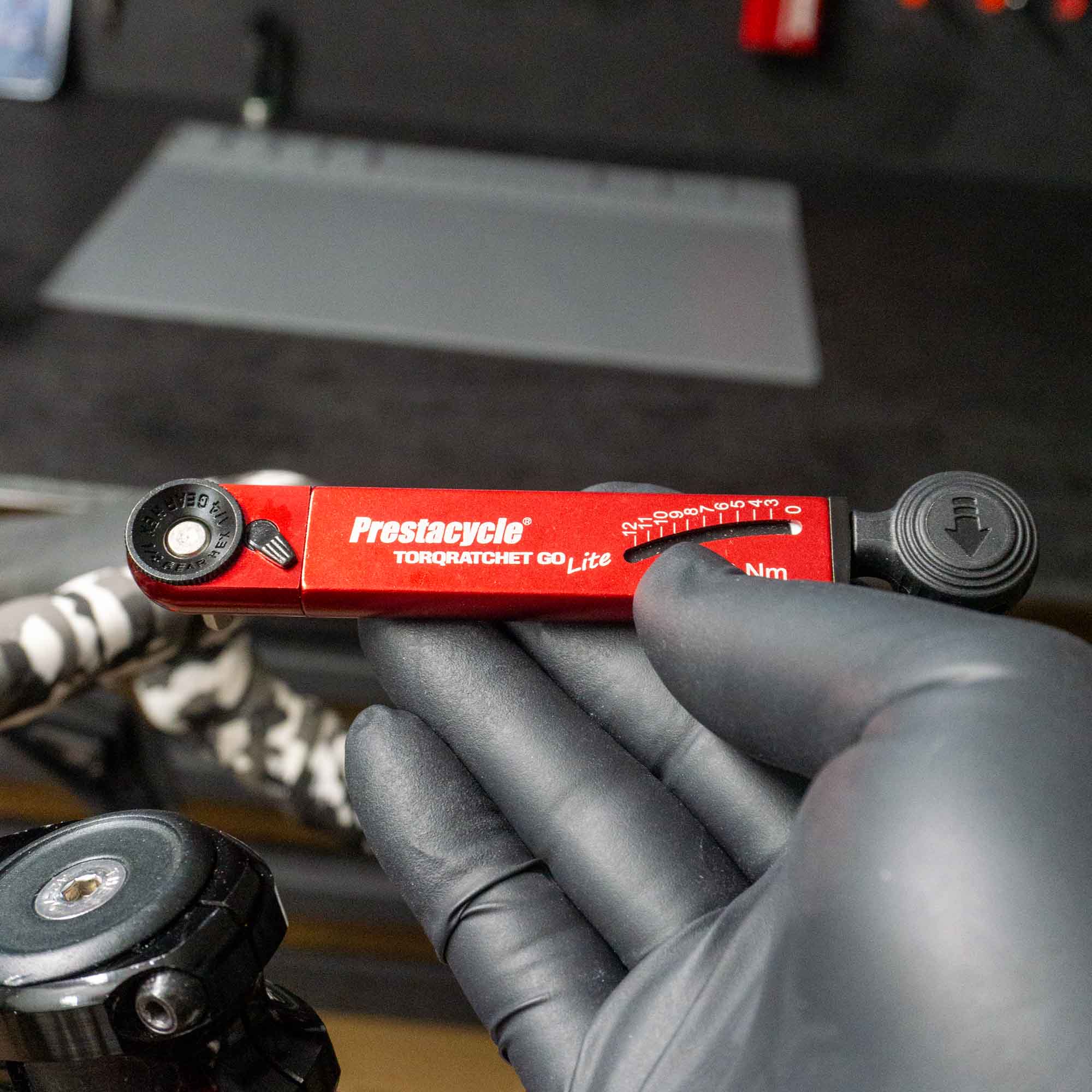 A gloved hand holds a red Prestacycle TorqRatchet GO Lite (3~12Nm) multi-tool ratchet, with a workbench, tools, and bike parts visible in the blurred background.
