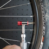 A close-up of a Prestacycle Prestaflator Mini Pro inflating a bicycle tire with its push-to-inflate Presta/Schrader valve. The red and silver tool stands out against a black, dotted background.