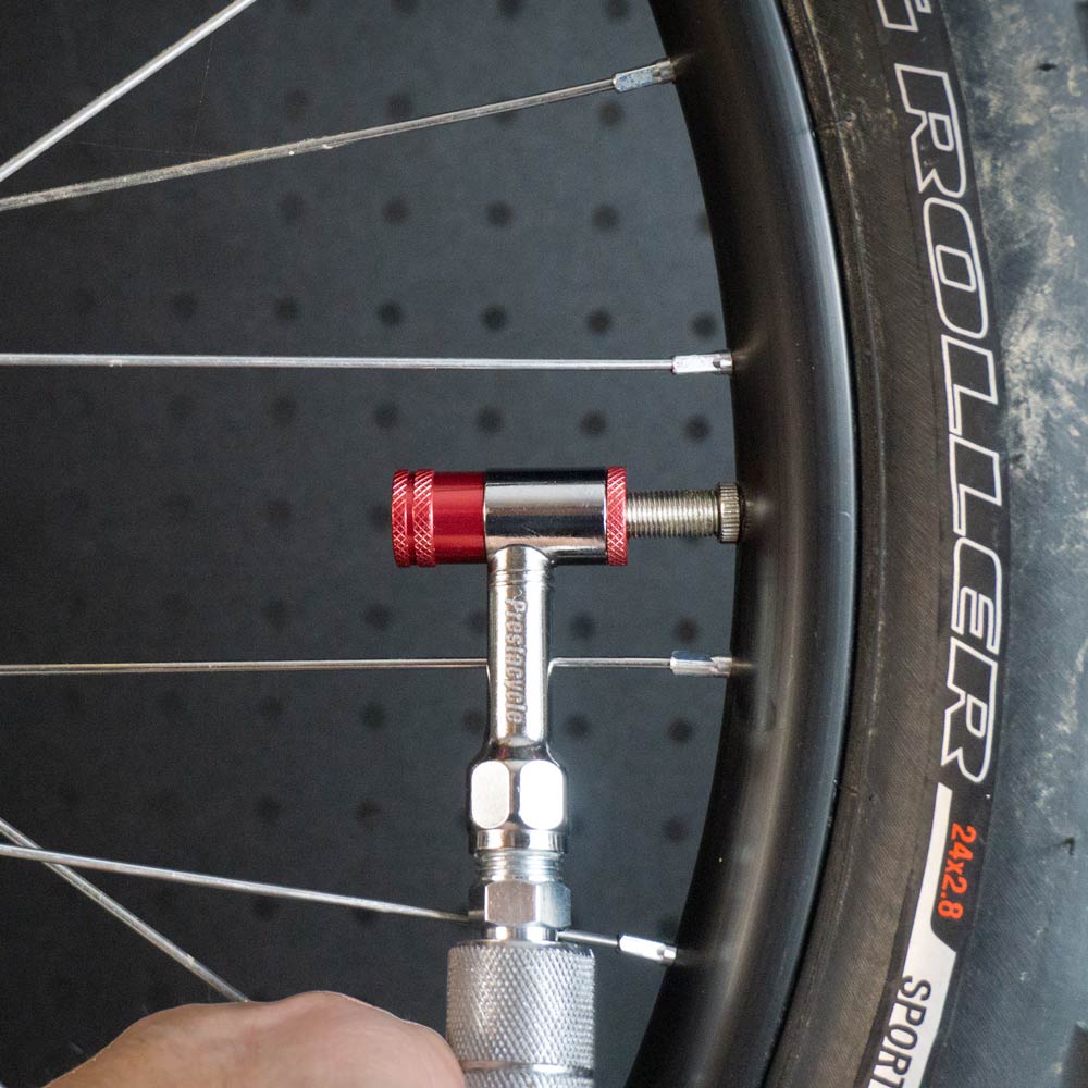 A close-up of a Prestacycle Prestaflator Mini Pro inflating a bicycle tire with its push-to-inflate Presta/Schrader valve. The red and silver tool stands out against a black, dotted background.