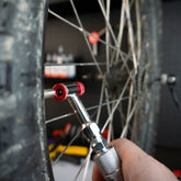 A close-up of a hand using the Prestacycle Prestaflator Mini Pro Bicycle Inflation Tool to check air pressure on a bike tire, highlighting its silver body, red fitting, cycling valve, and the bike wheel’s spokes and tread.
