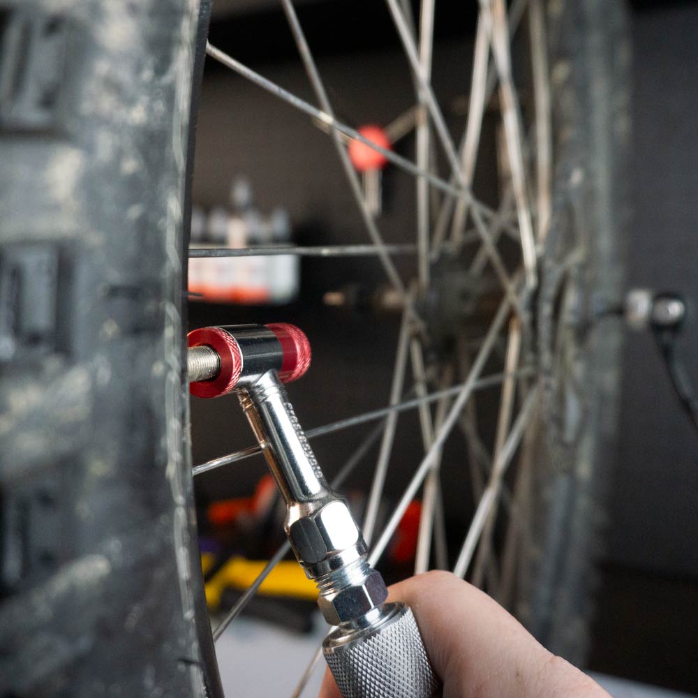 A close-up shows a hand using the Prestacycle Prestaflator Mini Pro – Presta / Schrader push-to-inflate Bicycle Inflation Tool to check air pressure, with the bike’s wheel, spokes, and valve clearly visible.