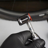 A gloved hand holds the Prestacycle Pro Presta Head near a bicycle tire, about to use its auto-lock feature for maintenance or inflation.