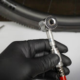 A gloved hand holds a Prestacycle Prestaflator PRO Bicycle Tire Inflator—Presta & Schrader Air Compressor Tool, attached to a red hose, near a bicycle wheel in the background.