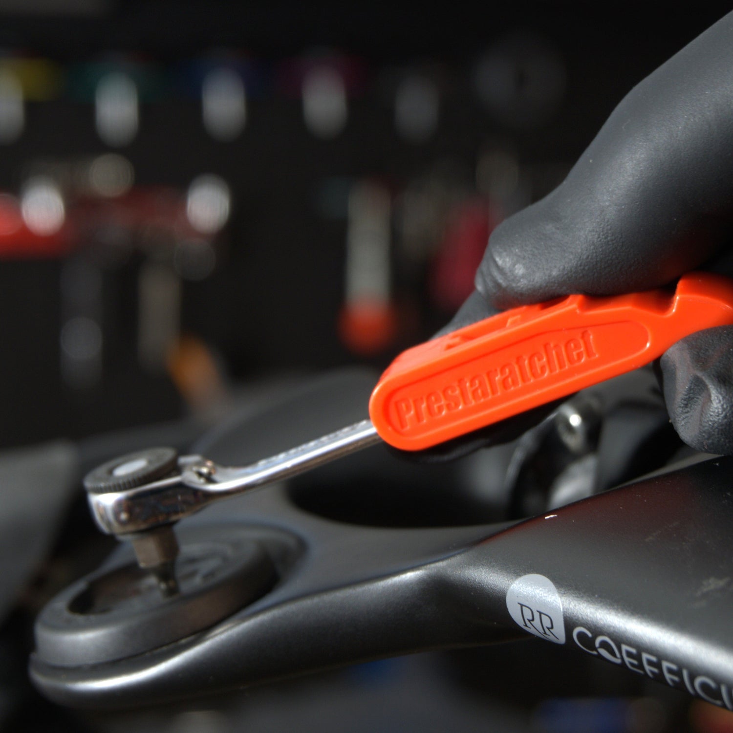 A gloved hand uses the Prestacycle Prestaratchet GO Multi-tool Kit to tighten a bolt on a dark-colored bike frame, with various tools blurred in the background.