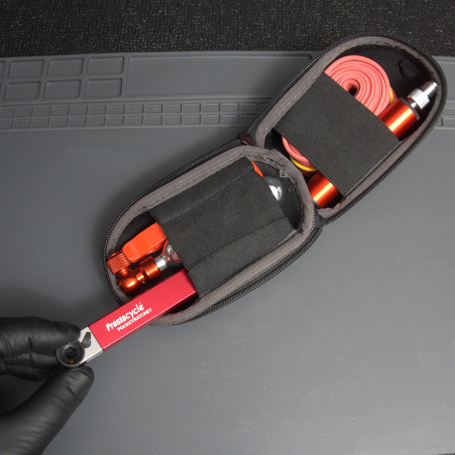 A gloved hand holds a compact zippered case containing a red Prestacycle PocketRatchet multi-tool with bits stored in the handle, a CO2 cartridge, a pink bicycle inner tube, and other essentials, all secured with elastic straps.