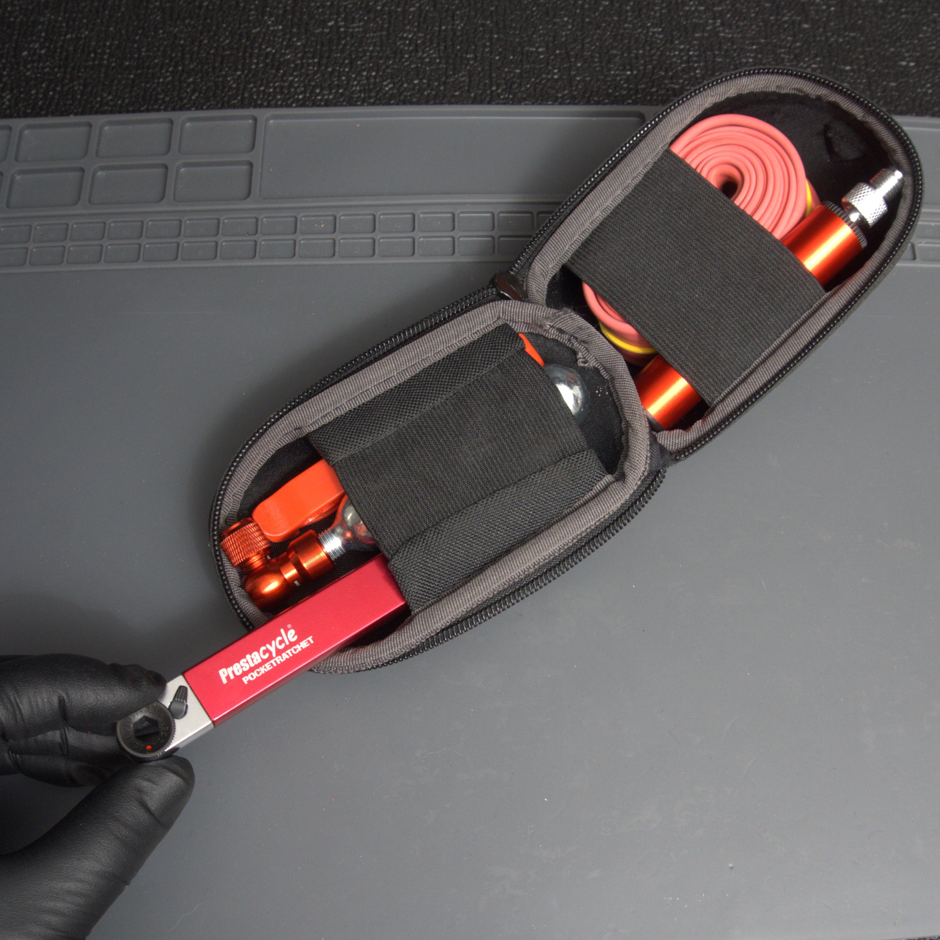 A gloved hand holds a compact zippered case containing a red Prestacycle PocketRatchet multi-tool with bits stored in the handle, a CO2 cartridge, a pink bicycle inner tube, and other essentials, all secured with elastic straps.